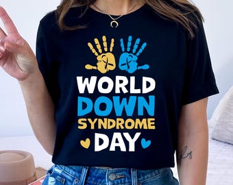 Pode incluir: Camiseta preta com um gráfico de duas impressões de mãos amarelas e azuis com uma fita no centro. O texto "WORLD DOWN SYNDROME DAY" é impresso abaixo das impressões de mãos.