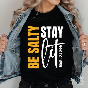 以下が含まれることがあります： 白い文字で「Be Salty Stay Lit」と「Matt. 5:13-14」と書かれた黒いTシャツ。「Be Salty」の文字は黄色です。