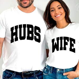May include: A white t-shirt with the word "HUBS" printed in black block letters. A white t-shirt with the word "WIFE" printed in black block letters.