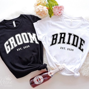 May include: Black and white t-shirts with the words "GROOM" and "BRIDE" printed in a bold, arched font. The text "EST. 2026" is below each word. The shirts are likely for a wedding or engagement celebration.