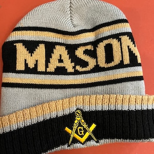May include: A gray knit beanie with the word "MASON" in gold letters on a black stripe. The hat features a gold and black striped brim with a Masonic emblem.
