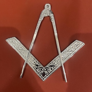 May include: A silver-colored Masonic square and compass emblem. The square and compass are intricately detailed with decorative patterns. The emblem is set against a red background.