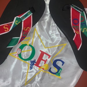 May include: A pair of black flip-flops with red, green, and white striped straps. The straps have the word "OFS" printed on them. The flip-flops are in a white satin drawstring bag with a yellow star and the word "OFS" printed on it.