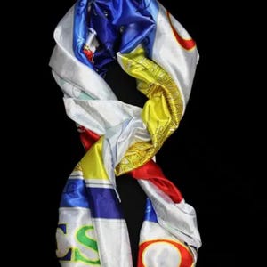 Puede incluir: Bufanda de seda colorida con un diseño vibrante. La bufanda presenta una mezcla de colores que incluyen blanco, azul, amarillo y rojo. La bufanda está drapeada sobre un fondo negro, resaltando sus intrincados patrones y texturas.