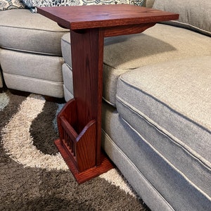 Pode incluir: Uma mesa de apoio de madeira de cor vermelha escura com um porta-revistas. A mesa é projetada para caber sobre o braço de um sofá.