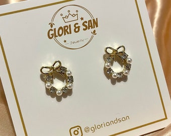 May include: A pair of gold-tone earrings featuring a small wreath design with faux pearls and rhinestones. The earrings are displayed on a white card with the text "GLORI & SAN - Jewelry - @gloriandsan".