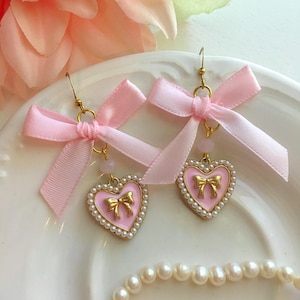 May include: A pair of pink heart-shaped earrings with gold trim and a gold bow in the center. The earrings are adorned with faux pearls and topped with pink ribbon bows. The earrings are displayed on a white plate.