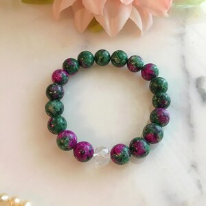 Puede incluir: Un brazalete de cuentas con cuentas de piedras preciosas verdes y magenta. Una cuenta de cristal transparente en el centro. Una flor rosa en el fondo. Joyería.