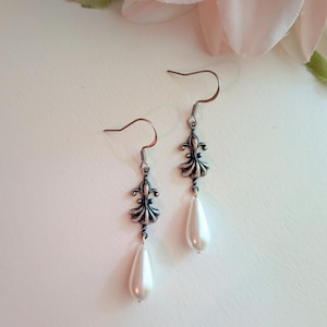 May include: A pair of drop earrings featuring a silver-toned metal fleur-de-lis connector attached to a hook wire. Each earring is finished with a smooth, white, teardrop-shaped faux pearl bead dangling from the bottom of the metal ornament.