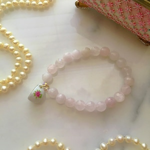 May include: A pink rose quartz bracelet with a small porcelain charm featuring a pink rose. The bracelet is made of faceted beads and is strung on a gold chain.