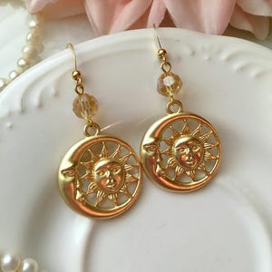May include: Gold-toned celestial earrings featuring a sun and moon design. Each earring has a sun face within a crescent moon, with a dangling amber-colored bead. The earrings are on a white plate, with a pearl necklace and pink flower in the background.