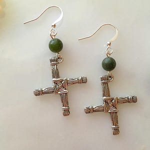 May include: A pair of silver-toned earrings featuring a St. Brigid's cross design. Each earring has a green bead and a hook closure. The crosses have a woven texture, and the earrings are displayed on a light-colored surface.