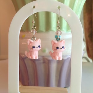 May include: Pink cat earrings with silver hooks. Each earring features a small, pink cat charm with black whiskers and blue eyes, accented with clear and blue beads. The earrings are displayed on a white stand.