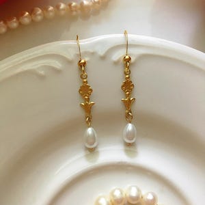 Puede incluir: Un par de pendientes de oro con una gota de perla y un diseño floral. Los pendientes están sobre una superficie blanca.