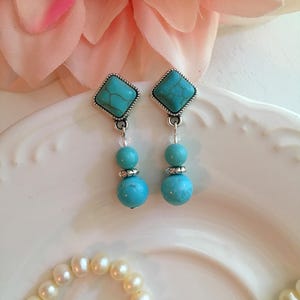 May include: A pair of turquoise earrings with silver-colored square tops and three turquoise beads. The earrings are displayed on a white plate with a pearl necklace and a pink flower in the background.