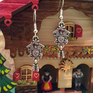 May include: Silver earrings with a cuckoo clock charm. The clock face has black Roman numerals and a black hand. The charm is attached to a silver chain with two silver bells.