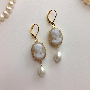 Peut inclure: Boucles d'oreilles dorées avec des pendentifs ovales en camée représentant des profils blancs et des perles en forme de goutte. Les boucles d'oreilles sont présentées sur une surface blanche, avec un collier de perles en arrière-plan.