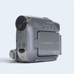 Puede incluir: Una videocámara digital Sony Handycam gris con lente Carl Zeiss Vario-Tessar y zoom óptico de 12x. El dispositivo incluye una pantalla LCD ancha montada en el lateral, una rejilla de altavoz y una correa de muñeca gris en el lateral.