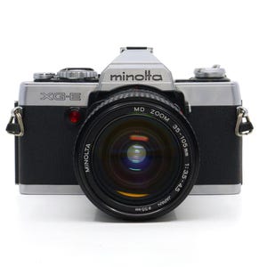 Puede incluir: Una cámara Minolta XG-E vintage con un objetivo zoom. El cuerpo de la cámara es negro y plateado, con el logotipo de Minolta. La lente es un MD Zoom 35-105mm con un diámetro de filtro de 55 mm.