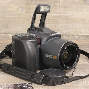 Pode incluir: Uma câmera Olympus preta com flash pop-up e tampa da lente. A câmera tem uma alça preta e a lente tem o texto "28-110 4X". A câmera está em uma superfície de madeira.