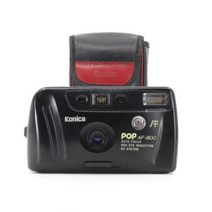 Könnte beinhalten: Eine schwarze Konica Pop AF-800 35mm Filmkamera mit eingebautem Blitz und einem System zur Reduzierung von Rote-Augen-Effekten. Die Kamera befindet sich in einem schwarzen Lederetui mit roter Innenausstattung.