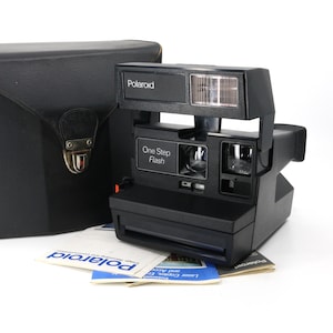 Peut inclure: Un appareil photo Polaroid One Step Flash noir avec un flash intégré et un étui de transport en cuir noir. L'appareil photo porte une étiquette indiquant "One Step Flash".