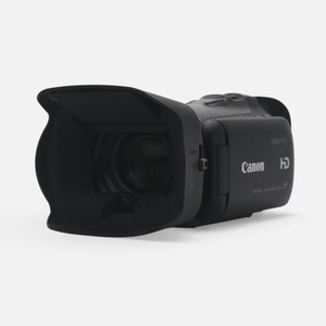 Op de afbeelding: Een zwarte Canon HD-camcorder met een grote zonnekap aan de voorzijde. Het apparaat heeft een compact, horizontaal ontwerp met het Canon-logo en de HD-aanduiding duidelijk zichtbaar op het zijpaneel bij de lensbehuizing.