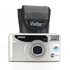 Op de afbeelding: Een zilveren Konica Z-up 125E camera met een Vivitar lens en een zwarte draagtas. De camera heeft een zoomlens met een bereik van 38-128 mm.