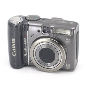 May include: A silver Canon PowerShot A590 digital camera with a 4x optical zoom lens and 8.0 megapixels. The camera has a black lens cap and a silver body with a black Canon logo.