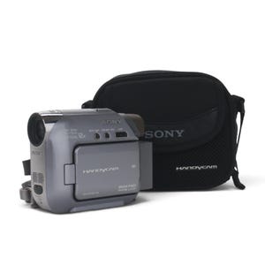 Op de afbeelding: Een zilverkleurige Sony Handycam digitale videocamera met Carl Zeiss-lens en 12x optische zoom, voorzien van een zijwaarts LCD-scherm met de tekst Handycam en Mega Pixel Wide LCD, vergezeld door een zwarte Sony-draagtas.