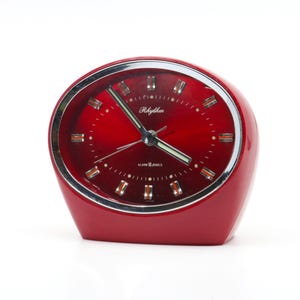 Könnte beinhalten: Ein roter Vintage-Wecker von Rhythm mit silberfarbener Lünette. Das Zifferblatt ist rot mit silberfarbenen Stundenmarkierungen und Zeigern. Die Uhr hat das Rhythm-Logo und die Worte "ALARM 2 JEWELS" auf dem Zifferblatt aufgedruckt.