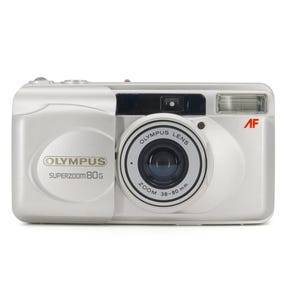 Op de afbeelding: Een zilveren Olympus Superzoom 80G camera met een zwarte lens en een ingebouwde flitser. De lens heeft de tekst "Olympus Lens Zoom 38-80 mm" erop gedrukt.
