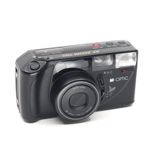 Może przedstawiać: Czarny aparat kompaktowy z obiektywem zoom 35-70 mm. Aparat ma wbudowaną lampę błyskową i samowyzwalacz. Aparat jest oznaczony jako "AF Zoom 700" i "M-Optic".