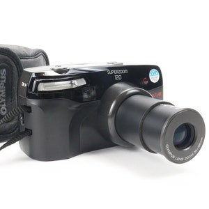 May include: A black Olympus Superzoom 120 camera with a lens that extends out. The camera has a black leather case with the Olympus logo on it.
