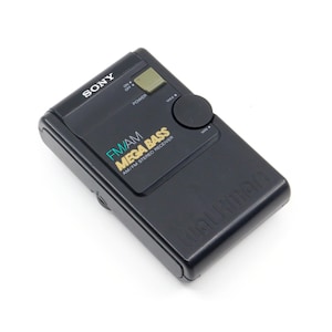 May include: A black Sony Walkman FM/AM radio with a green and blue "Mega Bass" logo. The radio has a power button, a volume knob, and a line in jack.