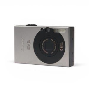 May include: A silver Canon Digital IXUS 70 compact camera with a black circular lens housing. The front features the text Canon Digital IXUS 70, 7.1 Mega Pixels, and lens specifications including 3x zoom, 5.8-17.4mm, and an aperture of 1:2.8-4.9.