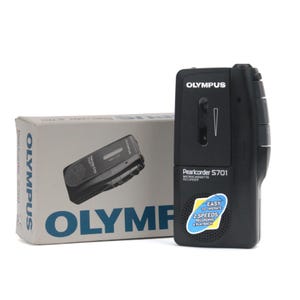 Op de afbeelding: Een zwarte Olympus Pearlcorder S701 microcassetterecorder met luidspreker en bedieningsknoppen. Het apparaat heeft een blauwe en gele sticker met de tekst Easy to Operate 2 Speeds Recording & Playback. Het staat naast de bijbehorende witte doos.