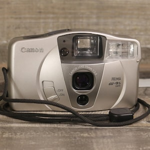 Op de afbeelding: Een zilveren Canon Prima AF-95 Date camera met een zwarte band. De camera heeft een Canon lens, een ingebouwde flitser en een datumfunctie.
