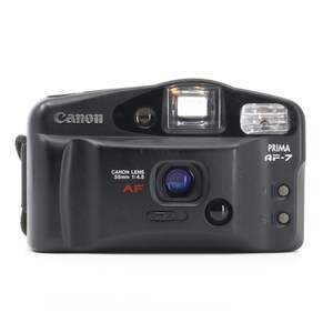 Op de afbeelding: Een zwarte Canon Prima AF-7 35mm filmcamera met een zilveren Canon-logo en rode tekst. De camera heeft een ingebouwde flitser en een lens met een 35mm 1:4,5 lens.
