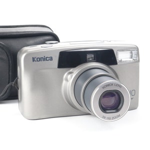 Op de afbeelding: Een zilveren Konica 38-110 zoomcamera met een zwarte leren tas. De camera heeft een zwarte lens met een zilveren ring eromheen. De camera heeft een zwart Konica-lenslabel op de lens.