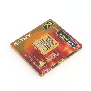 Puede incluir: Un MiniDisc Sony en un envoltorio de plástico transparente. El disco tiene un diseño degradado amarillo y naranja con las palabras "MiniDisc" en un cuadrado amarillo. También se ve el texto "topaz yellow" y "74".