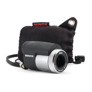 Puede incluir: Un microscopio de bolsillo Minox negro y plateado con una bolsa de transporte negra. El microscopio tiene una magnificación de 0,1 mm-∞ y una lente de 6x25.