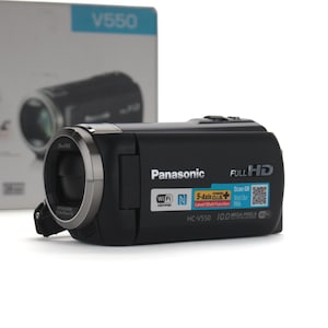 Puede incluir: Una videocámara Full HD Panasonic HC-V550 negra con un anillo de lente plateado. El cuerpo presenta etiquetas de Wi-Fi, NFC y 5-Axis Hybrid O.I.S. Plus, junto con un código QR y el texto 10.0 Mega Pixels. La caja del producto se ve detrás.
