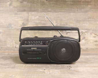 May include: Black vintage boombox with a handle and antenna. The radio cassette recorder has a speaker on the right side and a tuning dial. The text "AIWA" is visible on the front, along with "RM-21".