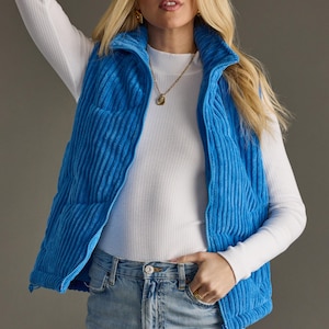 May include: A bright blue corduroy vest with a full zipper closure. The vest is worn over a white long-sleeve shirt and paired with light wash denim jeans. A gold necklace and ring complete the look.