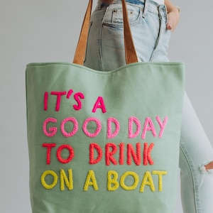 May include: A light green tote bag with brown leather handles. The bag features the embroidered phrase "IT'S A GOOD DAY TO DRINK ON A BOAT" in pink, red, and yellow thread. The bag is held by a person wearing light blue jeans.