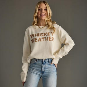 May include: A cream-colored long-sleeve knit sweater featuring the text WHISKEY WEATHER in brown block letters across the chest, paired with light blue denim jeans. The sweater has a ribbed crew neckline, ribbed cuffs, and a ribbed hemline.