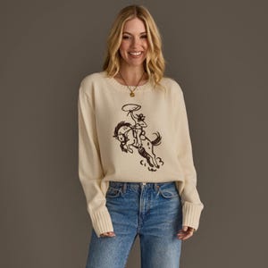 May include: A cream-colored long-sleeve knit sweater featuring a dark brown line-art graphic of a cowboy riding a bucking horse while swinging a lasso. The sweater has a ribbed crew neckline and wide ribbed cuffs on the sleeves.