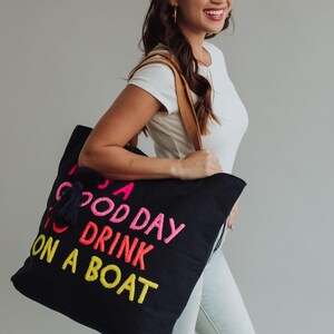 May include: A black tote bag with the embroidered phrase "A Good Day to Drink on a Boat" in pink, orange, and yellow. The bag has a navy blue tassel and brown leather straps. The woman is wearing a white t-shirt and light blue jeans.