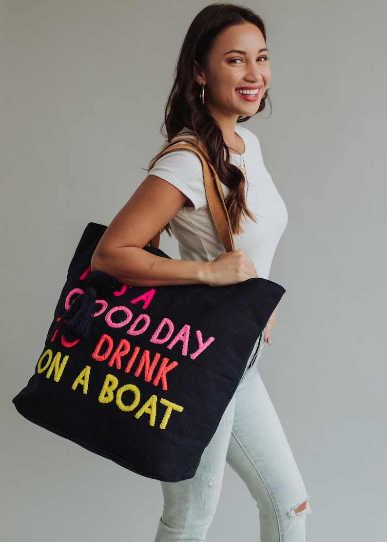 May include: A black tote bag with the embroidered phrase "A Good Day to Drink on a Boat" in pink, orange, and yellow. The bag has a navy blue tassel and brown leather straps. The woman is wearing a white t-shirt and light blue jeans.